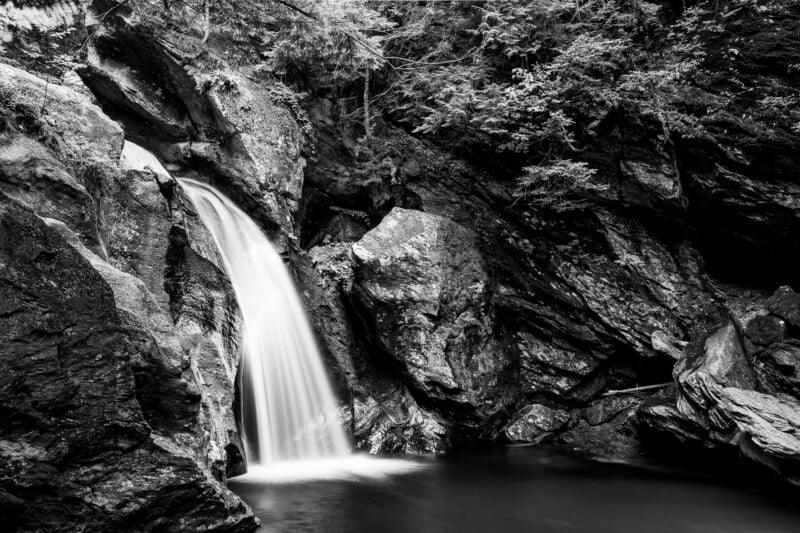 A black and white long-exposure shot of a waterfall cascading over dark, jagged rocks into a calm pool.