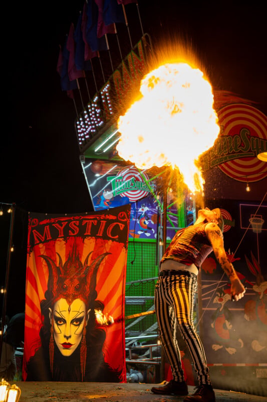 A performer in striped pants and a vest breathes a large fireball in front of a carnival game booth featuring a banner with a stylized portrait.