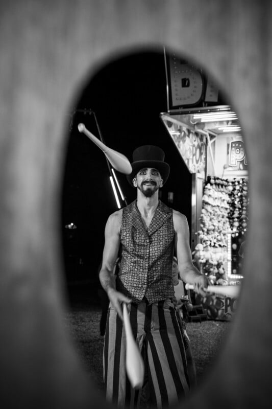 A man in a top hat and patterned vest juggles white clubs while framed by a dark, oval-shaped opening at a carnival.