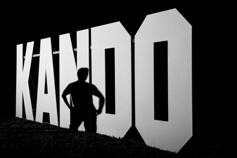 A black and white shot of a person's shadow cast against a large, white sign spelling "KANDO" at night.