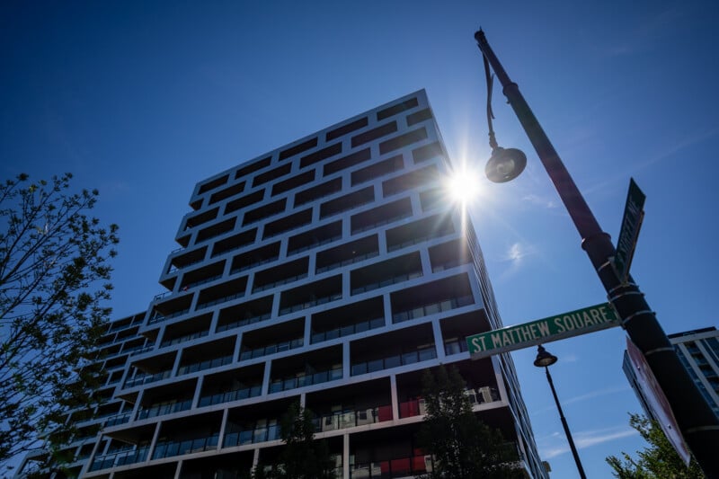 A modern, multi-story apartment building with a grid-like facade stands against a bright blue sky, with a street sign for St. Matthew Square in the foreground.