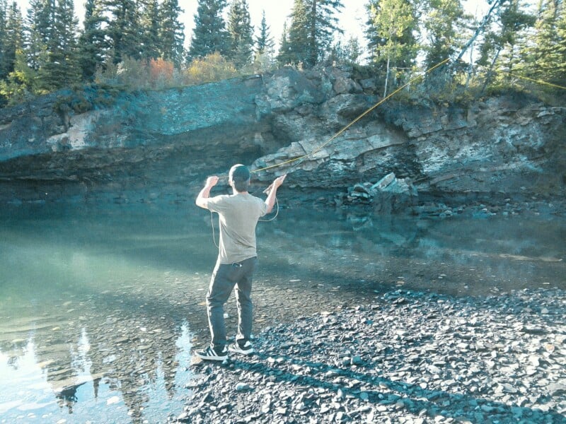 A person stands on a rocky shore fly fishing in a calm, clear river with a forested cliff in the background.
