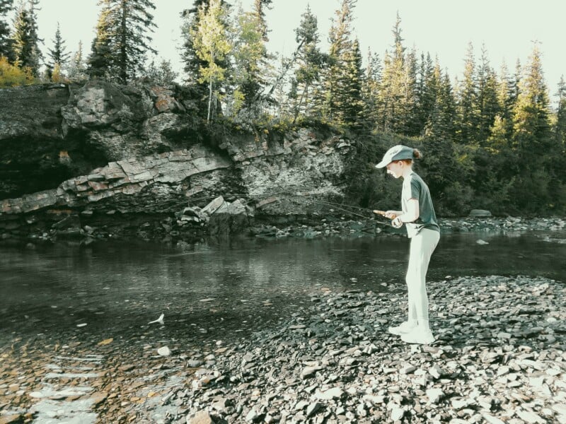 A person wearing a cap and light-colored clothing stands on a rocky riverbank, holding a fishing rod over the water.