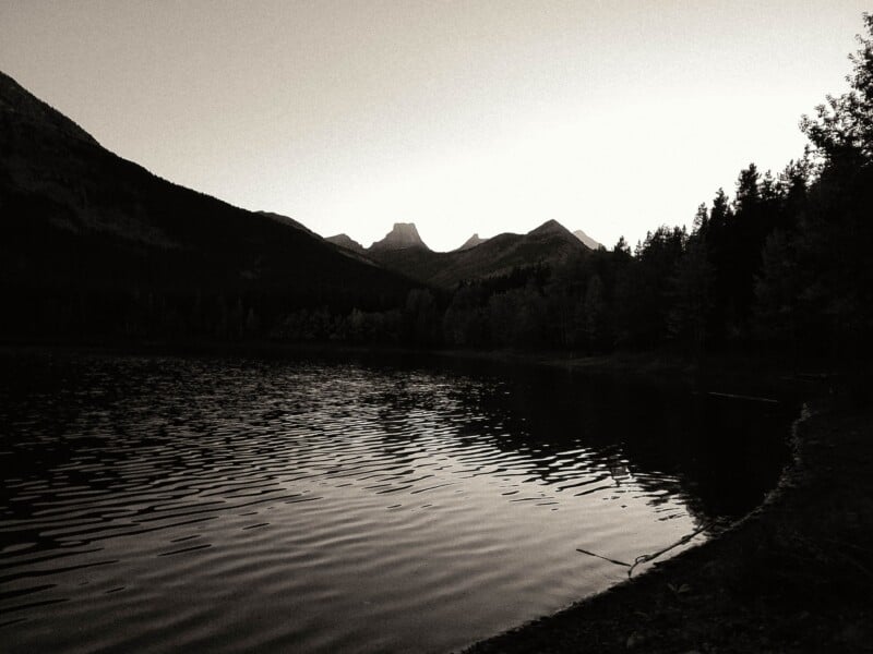 A black and white landscape of a calm lake reflecting the sky, framed by dark, silhouetted mountains and a forest shoreline.