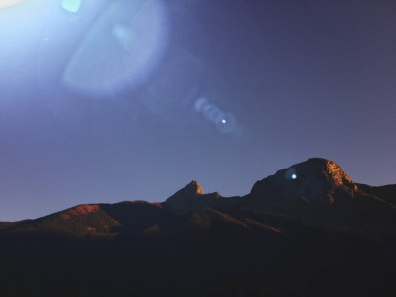 Sunlight illuminates the peaks of a dark mountain range against a clear, deep blue sky with lens flare.