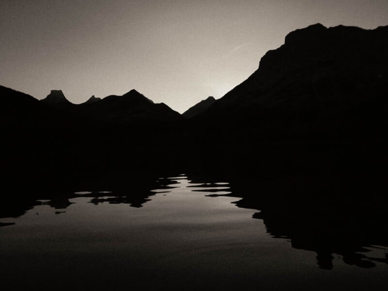 A high-contrast, monochromatic silhouette of jagged mountain peaks reflected in the calm, dark water of a lake.