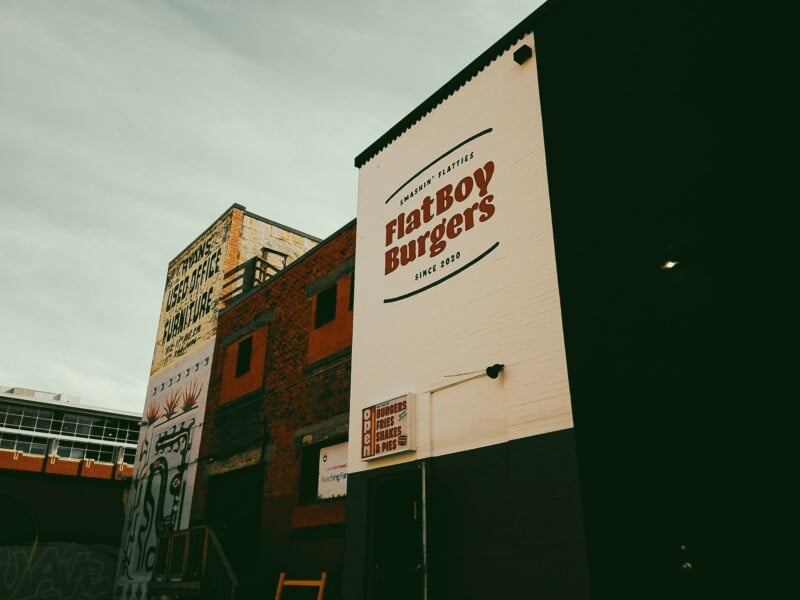A low-angle shot of a brick building with a large sign for FlatBoy Burgers painted on its white side wall, next to an older building with faded signage for used office furniture.