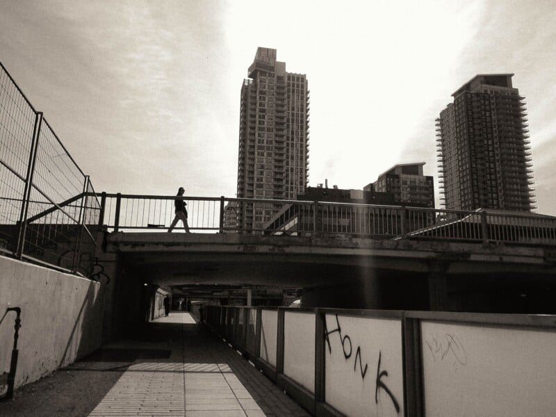 A person walks across a pedestrian bridge above a shadowed walkway with graffiti on a glass barrier, set against tall buildings.