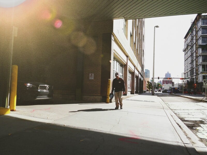A man in a dark sweatshirt and tan pants walks along a city sidewalk under an overpass, with a parked car to his left and a tall building on the right.