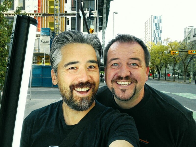 Two men with beards smile at the camera in a selfie taken on a city street near a construction site.