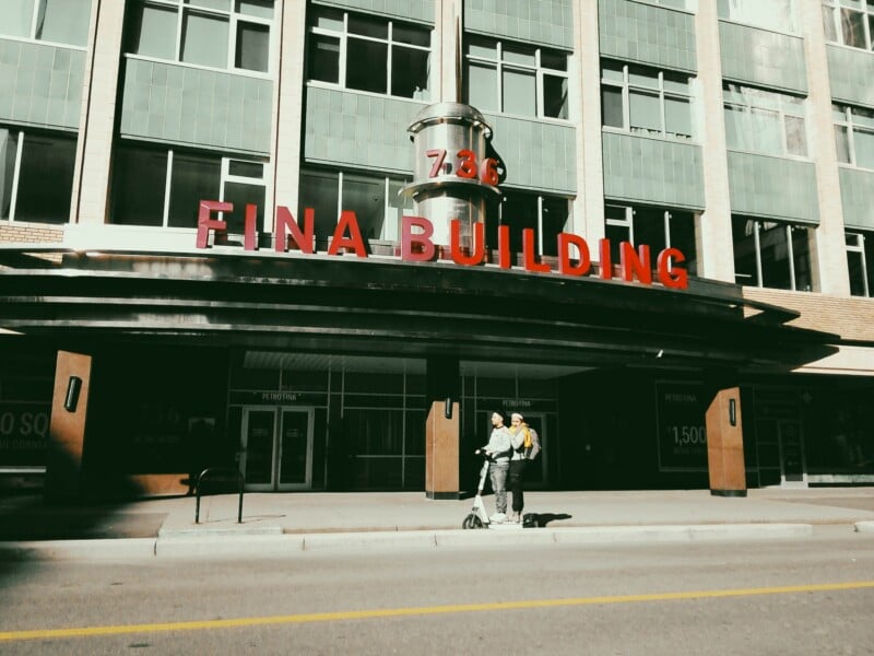Two people ride an electric scooter together on the sidewalk in front of the Petro Fina Building.