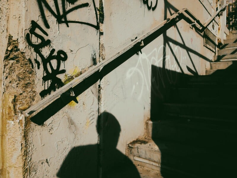 A concrete staircase with a metal handrail is shown next to a weathered wall covered in graffiti, with the shadow of a person cast onto the wall.