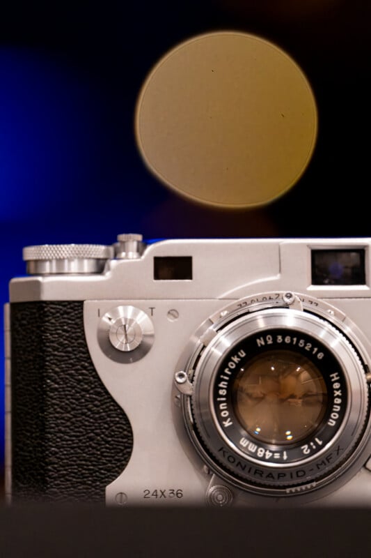A close-up of a vintage silver film camera with a Konishiroku Hexanon lens, set against a dark background with a large, out-of-focus yellow circle above it.