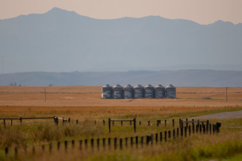 A row of seven metal grain silos stands in a golden field against a backdrop of distant mountains.