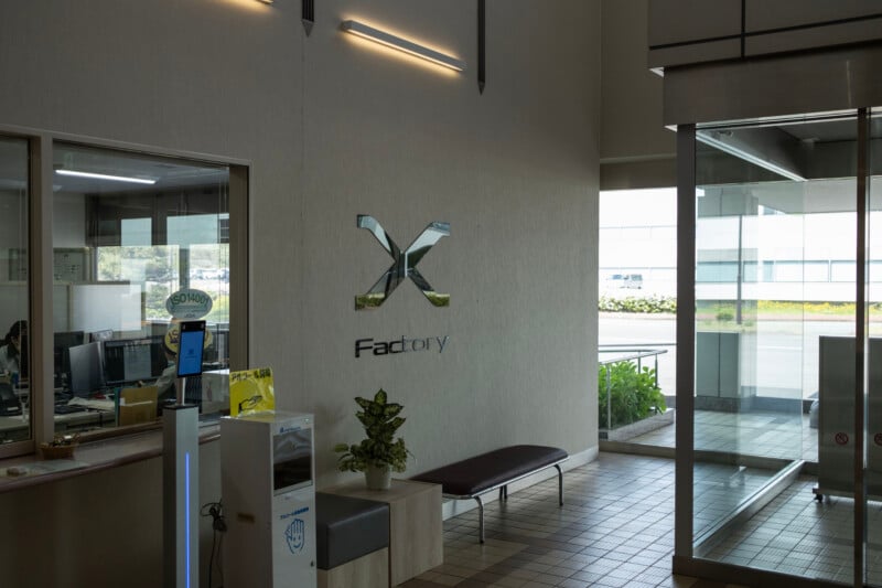 The interior of a building entrance features a wall with a large metallic "X" logo above the word "Factory," a hand sanitizer station, a small potted plant, and a bench near a glass door.