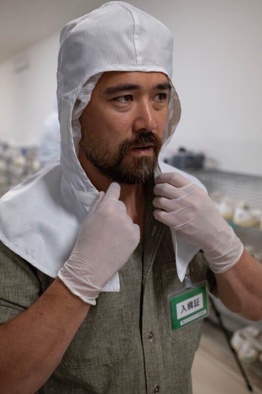 A man with a beard wearing a white cleanroom hood and gloves adjusts the fabric around his neck while wearing a green ID badge.