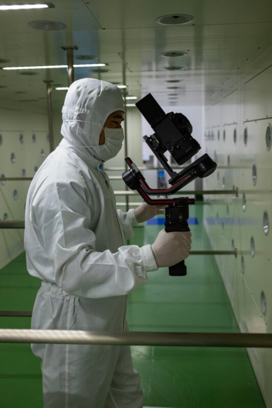 A person in a white cleanroom suit and face mask holds a camera mounted on a handheld gimbal inside a sterile facility.