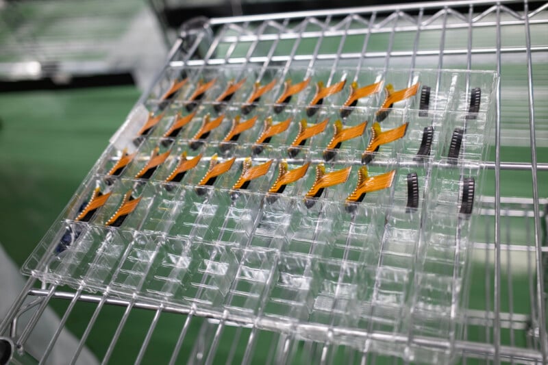 A plastic tray containing rows of orange flexible ribbon cables and black electronic components sits on a wire shelving rack.