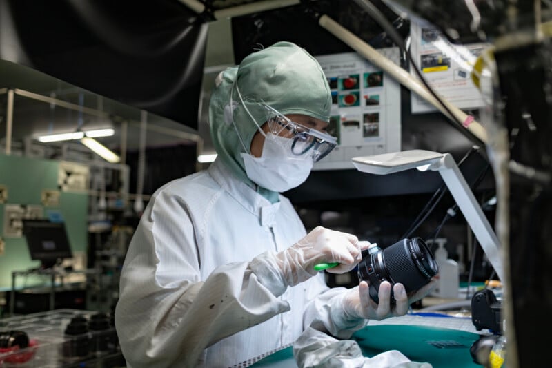 A person in a cleanroom suit, mask, and safety goggles cleans a camera lens with a small brush under a desk lamp.