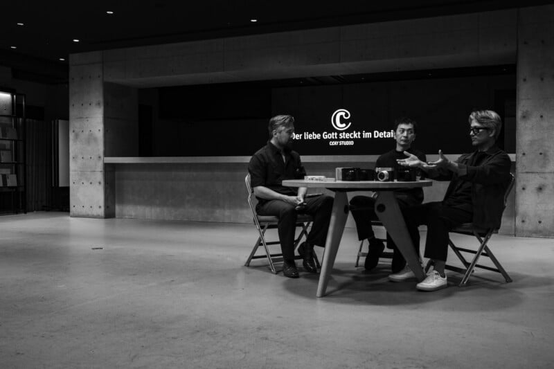 Three men sit around a small table in a minimalist, concrete-walled room, with a logo and text reading "Der liebe Gott steckt im Detail" on the wall behind them.