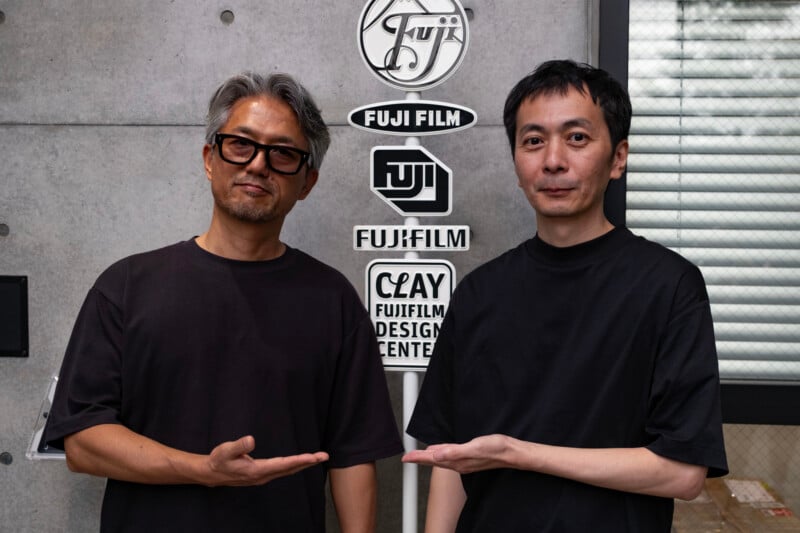 Two men in black t-shirts stand in front of a vertical sign displaying various Fujifilm logos and the text "CLAY FUJIFILM DESIGN CENTER," both gesturing toward the sign with their hands.