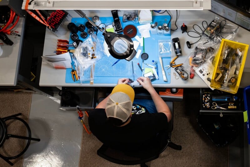 A person wearing a baseball cap sits at a workbench repairing camera equipment, surrounded by tools, lenses, and parts on a blue cutting mat.