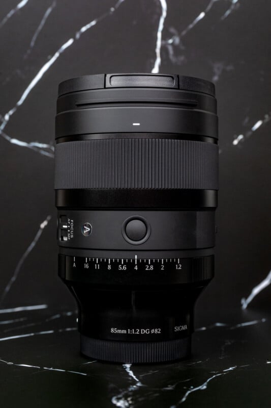 A black Sigma 85mm 1:1.2 DG Ø82 camera lens stands upright against a black and white marble-patterned background.