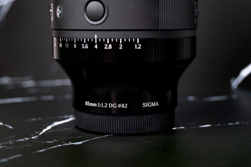 A close-up of a black camera lens standing upright on a black marble surface, showing aperture markings from 1.2 to 16 and text reading 85mm 1:1.2 DG 82 SIGMA.