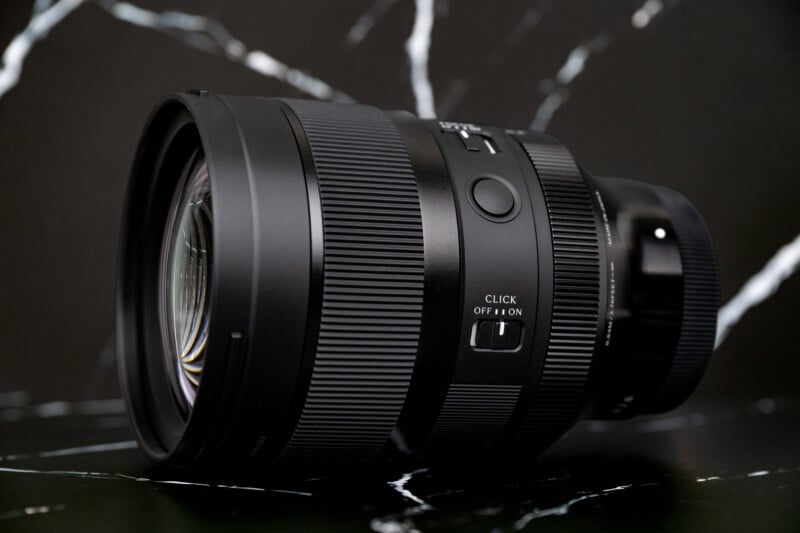 A black camera lens sits on a dark, marble-patterned surface, featuring a focus ring, a customizable button, and a switch labeled "CLICK OFF | ON.