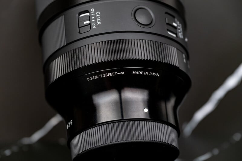 A close-up view of a black camera lens barrel featuring a focus ring, a click switch, and text indicating a minimum focus distance of 0.84M/2.76FEET and "MADE IN JAPAN.