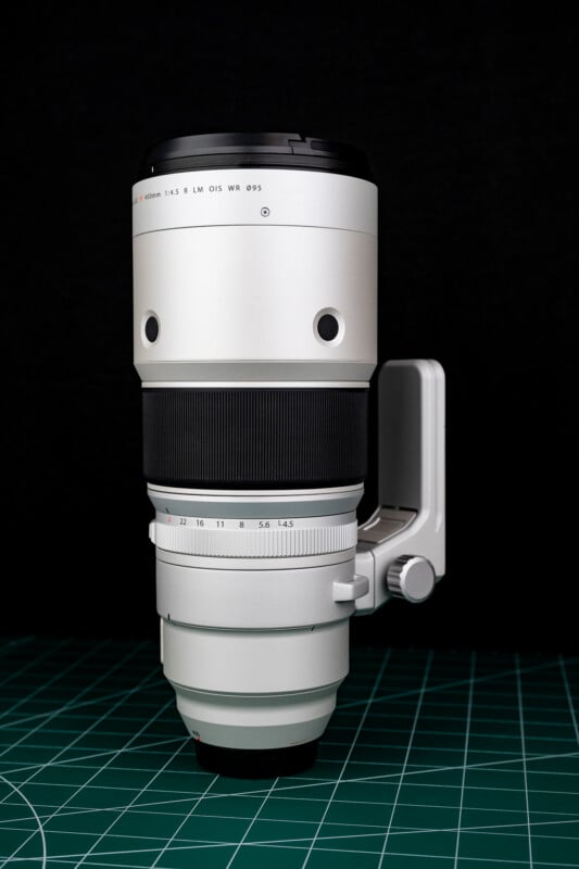 A silver telephoto camera lens stands vertically on a green cutting mat against a black background.