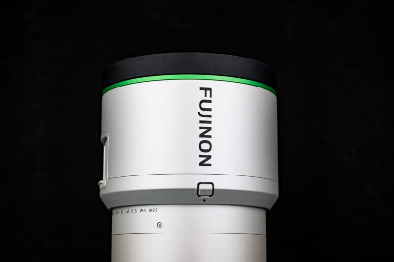 A close-up view of a silver Fujinon camera lens against a black background, featuring a green accent ring and a visible focus button.
