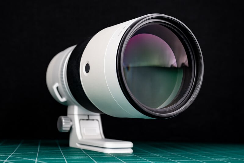 A white telephoto camera lens with a black focus ring sits on a green cutting mat against a black background.