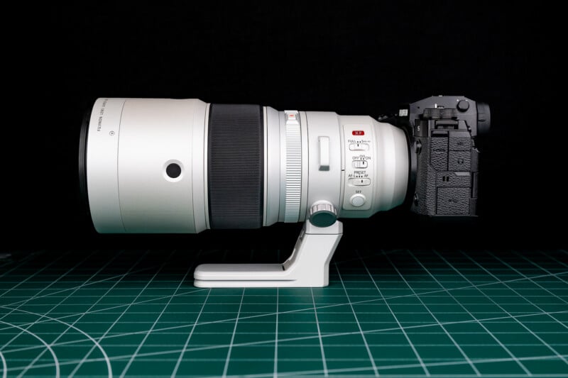 A white telephoto lens attached to a black mirrorless camera body, resting on a green cutting mat against a black background.