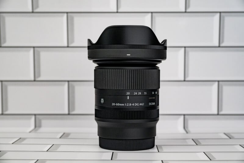 A black Sigma 20-60mm camera lens stands upright against a white subway tile background.