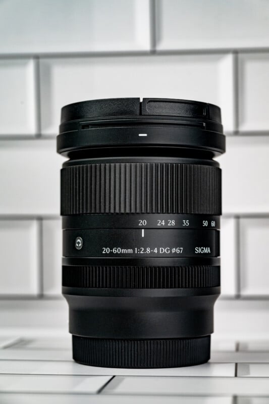 A black Sigma 20-60mm 1:2.8-4 DG 67mm camera lens stands upright against a white subway tile background.