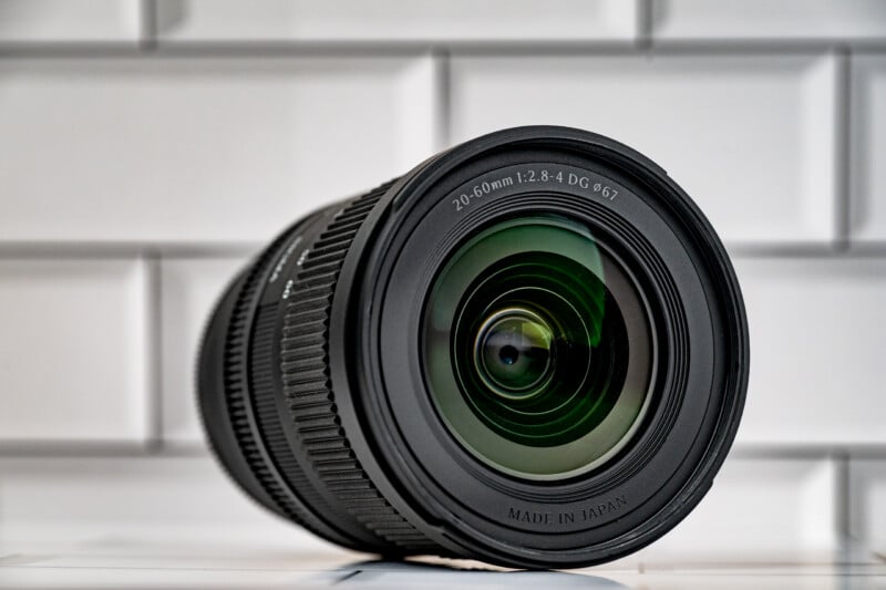 A black camera lens with a 20-60mm 1:2.8-4 DG 67mm specification sits on a white tiled surface against a white brick background.