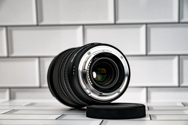 A camera lens rests on its side on a white tiled surface next to a black lens cap, showing its rear mount and electrical contacts.