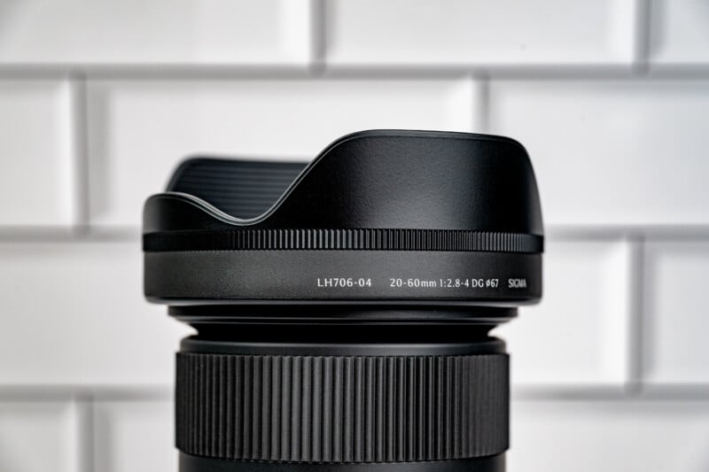 A close-up of a black camera lens hood attached to a lens, with text reading "LH706-04 20-60mm 1:2.8-4 DG ø67 SIGMA" against a white tiled background.