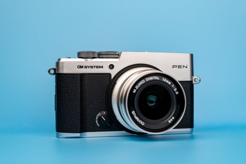 A silver and black OM SYSTEM PEN digital camera with a lens attached, centered against a solid light blue background.