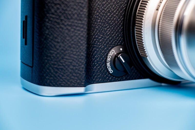 A close-up of a camera body featuring a physical switch labeled "COLOR/MONO" set to the color position, with a silver lens visible on the right against a light blue background.