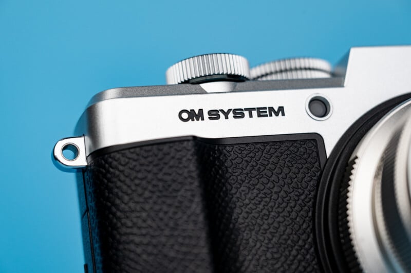 A close-up view of the top corner of a silver and black camera body with the text "OM SYSTEM" printed on the front.