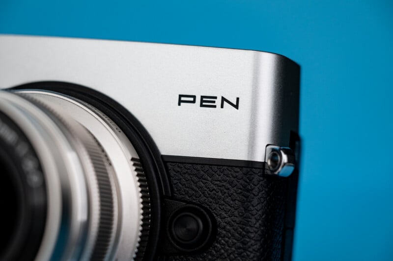 A close-up view of a silver and black camera body with the word "PEN" printed on the top panel against a solid blue background.