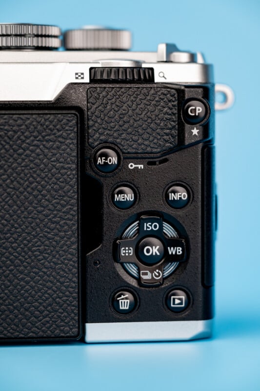 A close-up view of the back of a digital camera, showing buttons labeled AF-ON, MENU, INFO, OK, ISO, WB, a trash icon, and a playback icon against a blue background.