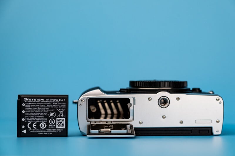 An OM SYSTEM BLS-7 camera battery sits next to a silver camera with its battery compartment door open, set against a blue background.
