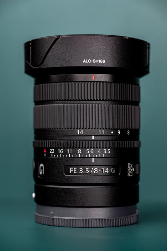 A black Sony FE 3.5/8-14 G camera lens stands vertically against a solid teal background, showing its zoom and aperture rings.