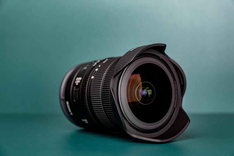 A black camera lens with a petal-shaped hood rests on a solid teal surface against a matching teal background.