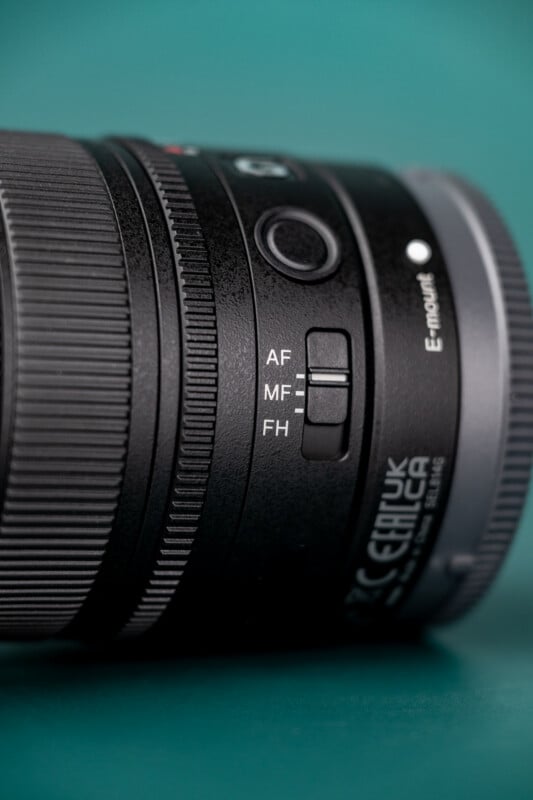 A close-up of a camera lens barrel featuring an E-mount label and a focus mode switch set to MF, positioned against a teal background.