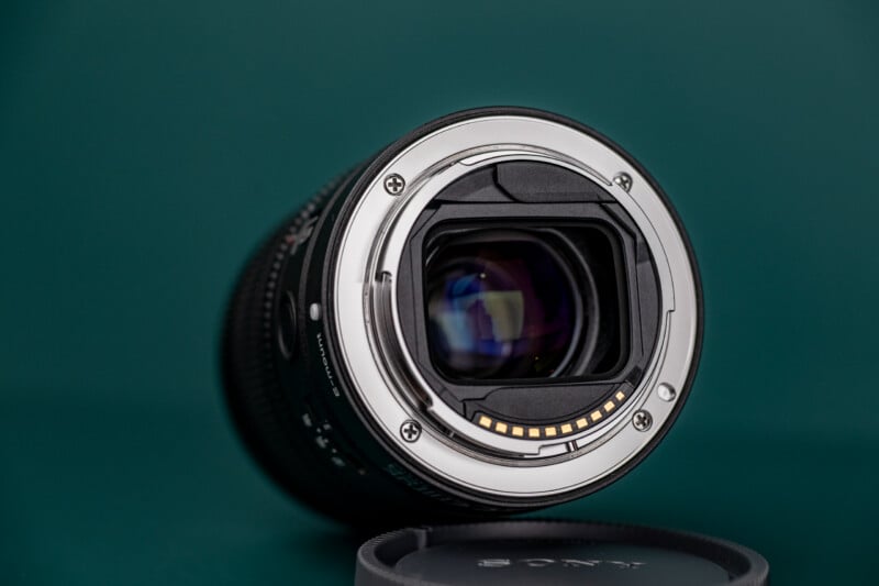A Sony E-mount camera lens is positioned on a dark green surface, showing its rear metal mount, electronic contact pins, and internal glass elements, with a lens cap lying in the foreground.