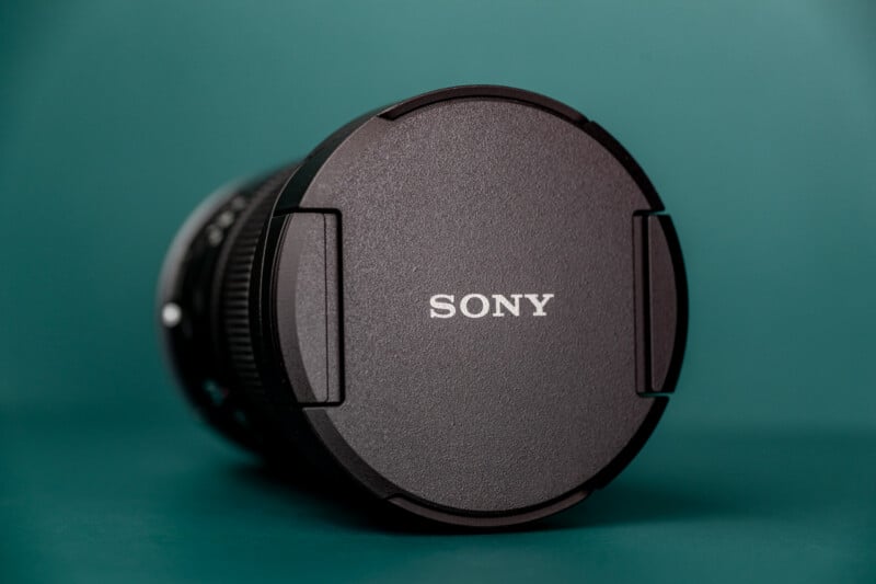 A black Sony camera lens cap sits on a teal surface, angled slightly to show the side of the lens barrel.